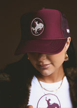 Load image into Gallery viewer, Person wearing a maroon trucker hat with a logo, standing against a dark background
