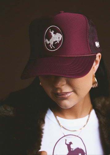 Person wearing a maroon trucker hat with a logo, standing against a dark background